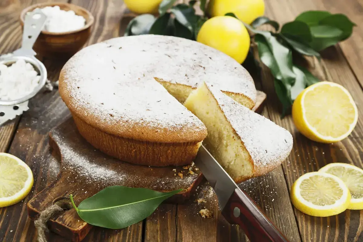 Una fetta di torta al limone morbidissima e profumatissima benedetta, cosparsa di zucchero a velo, con limoni freschi e foglie verdi su un tagliere di legno.
