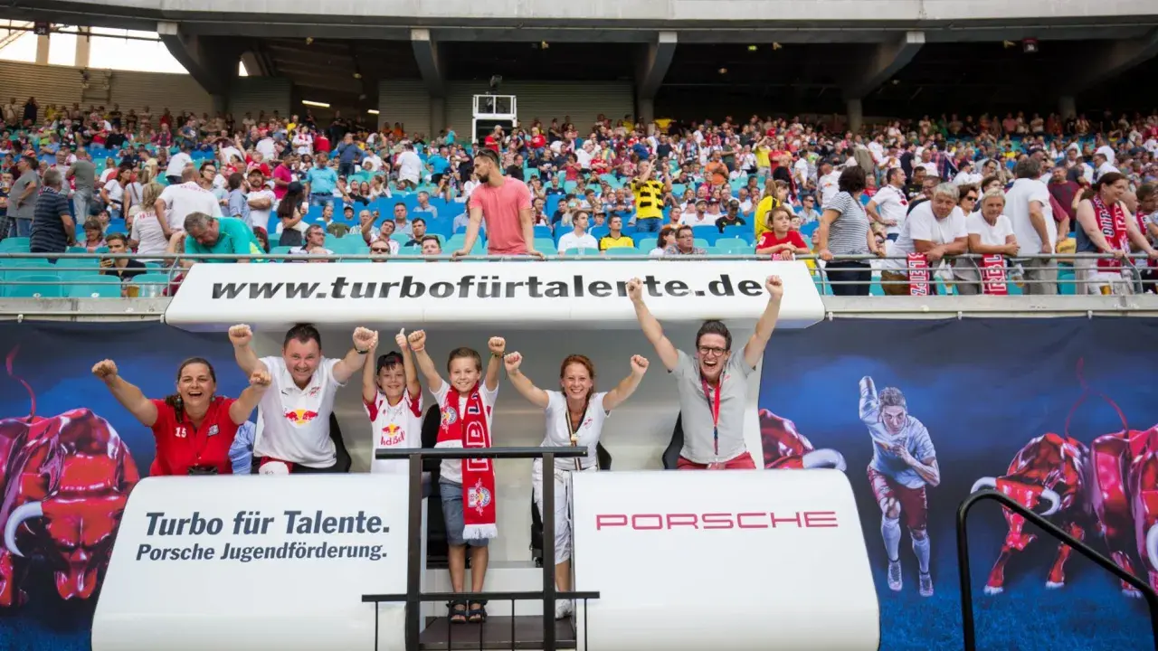 Zwei Kinder und zwei Erwachsene jubeln auf einer Tribüne vor einem Banner mit Bullen und einem Läufer.