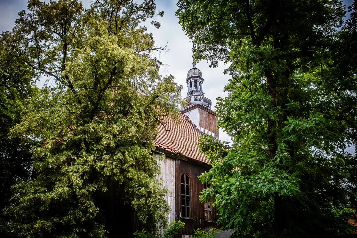 Stary kościół w Miejskiej Górce, otoczony bujną zielenią drzew, z charakterystyczną wieżą i drewnianą elewacją.