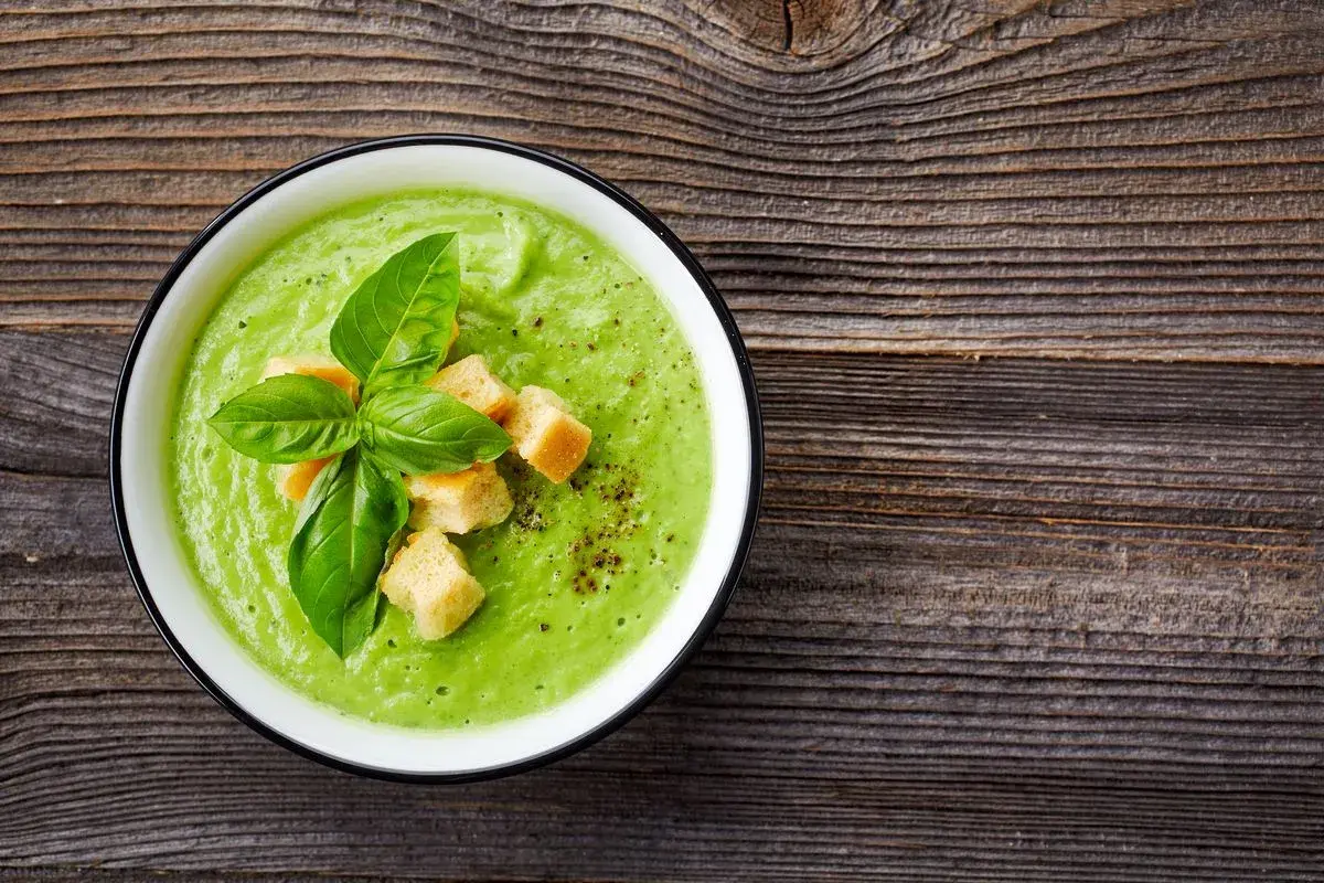 Un cremoso passato di verdure verde brillante, guarnito con crostini dorati e basilico fresco, su un tavolo di legno rustico.