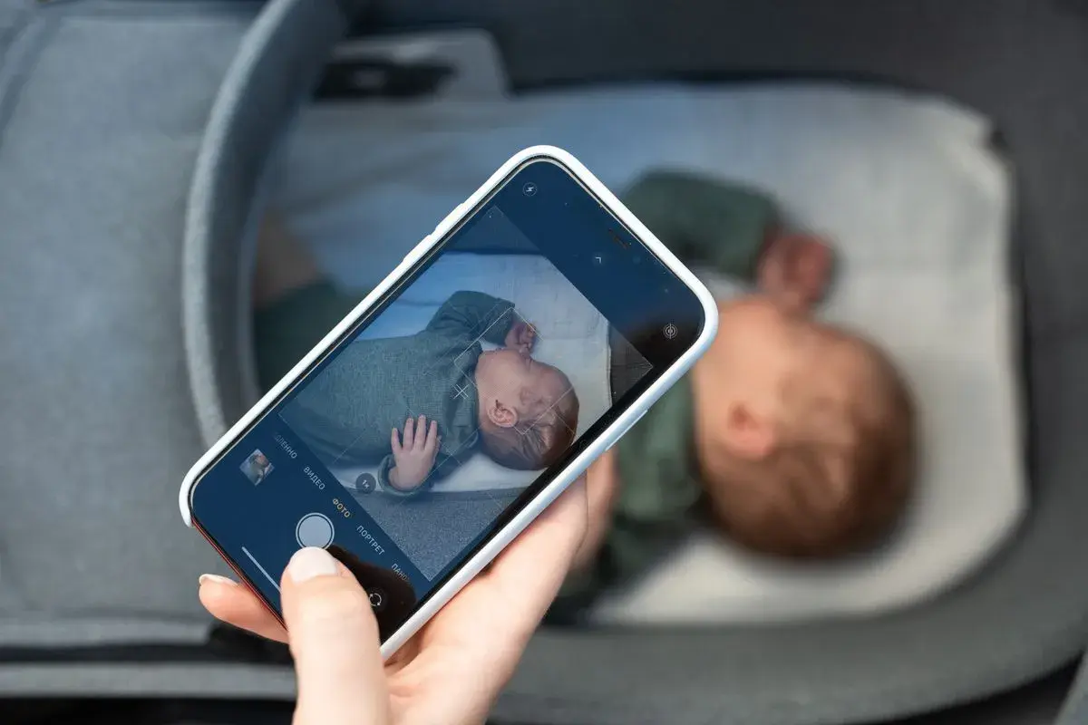 En hand håller en mobiltelefon och fotograferar en sovande bebis i en vagn. En billig babyvakt kan vara bra för att hålla koll.