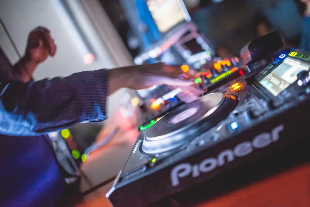 Les mains d'un DJ mixent sur une platine Pioneer, là où les DJs trouvent leur musique. Lumières colorées et ambiance festive.