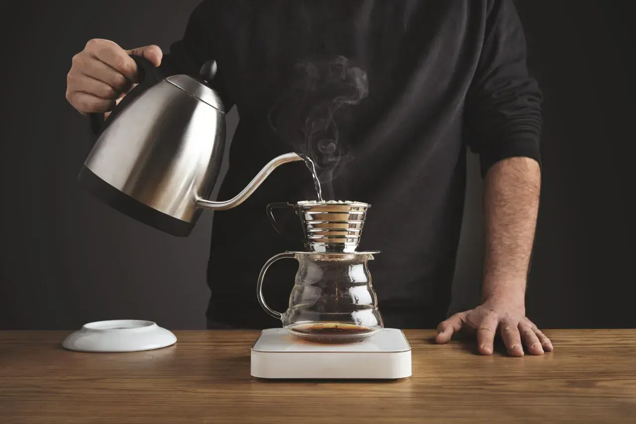 Mężczyzna przelewa gorącą wodę z czajniczka do drippera Kalita, przygotowując aromatyczną kawę.