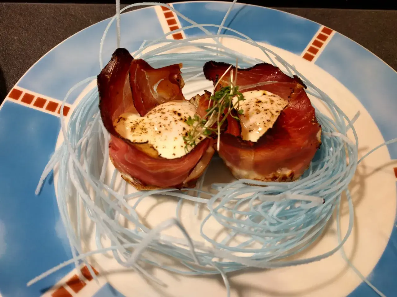 Zwei Eier mit Speck, kunstvoll in einem Nest aus blauen Fäden auf einem Teller angerichtet.