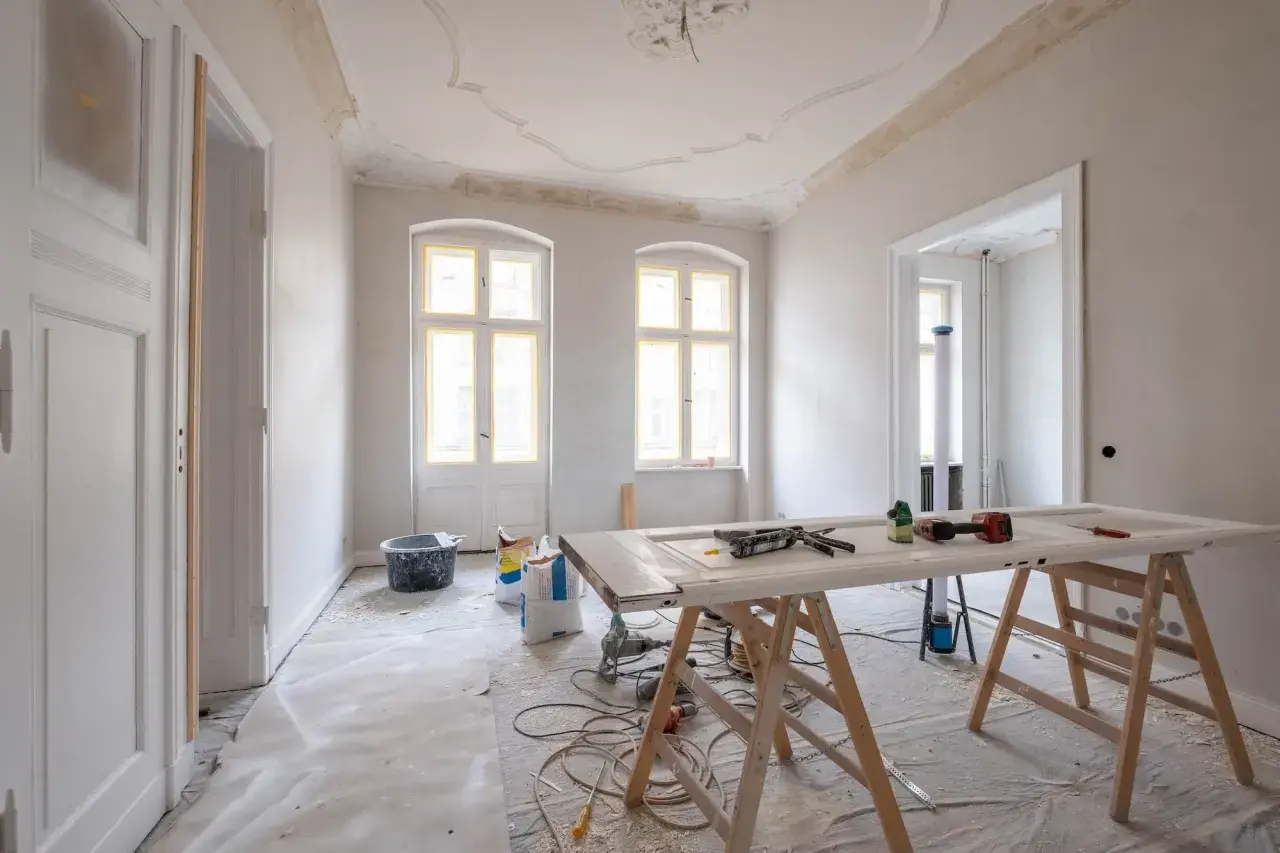 Mieszkanie na sprzedaż do remontu. Wnętrze w trakcie prac, widoczne materiały budowlane, drzwi na kozłach, okna i drzwi z żółtą taśmą.