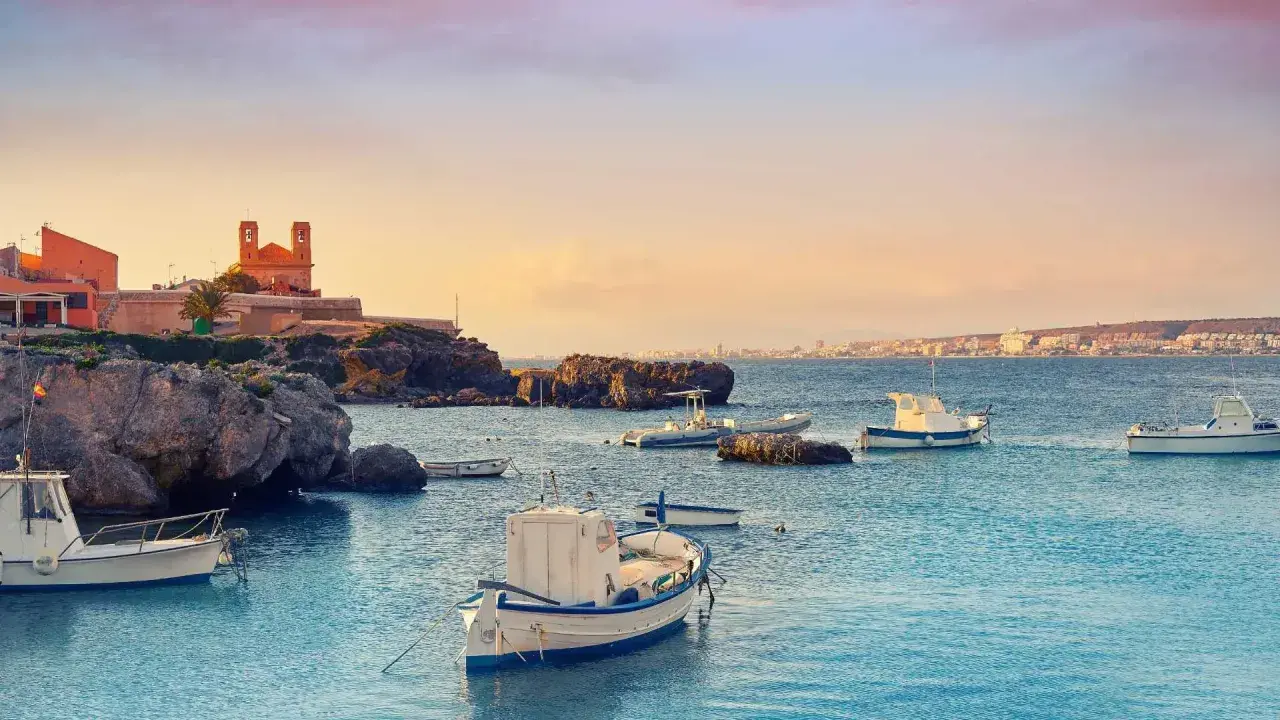 Barcas tranquilas en el puerto Santa Pola, con edificios históricos y la costa al fondo bajo un cielo anaranjado.