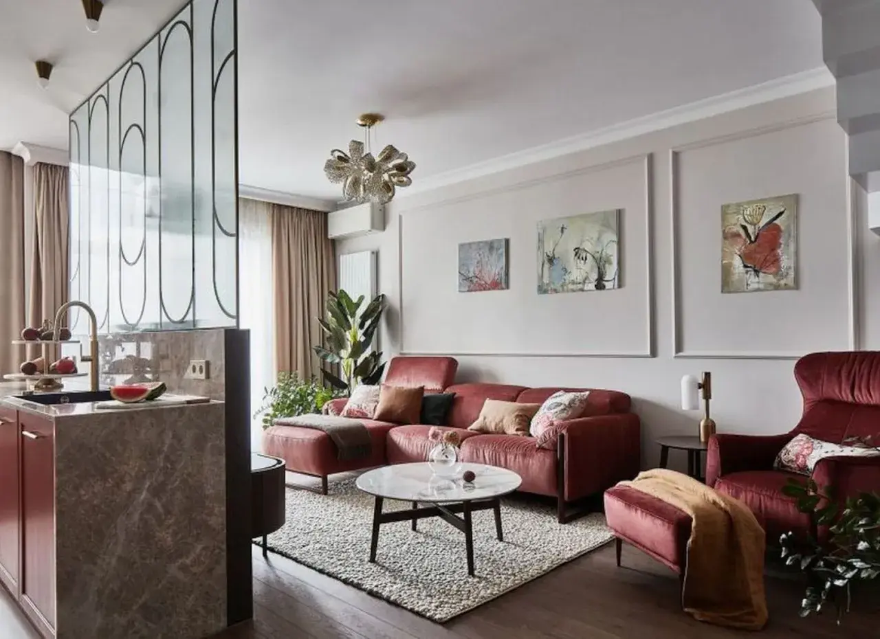 Elegancki salon z bordową kanapą i fotelem. Modne ściany w salonie zdobią obrazy, a kuchnia oddzielona jest ażurową ścianką.