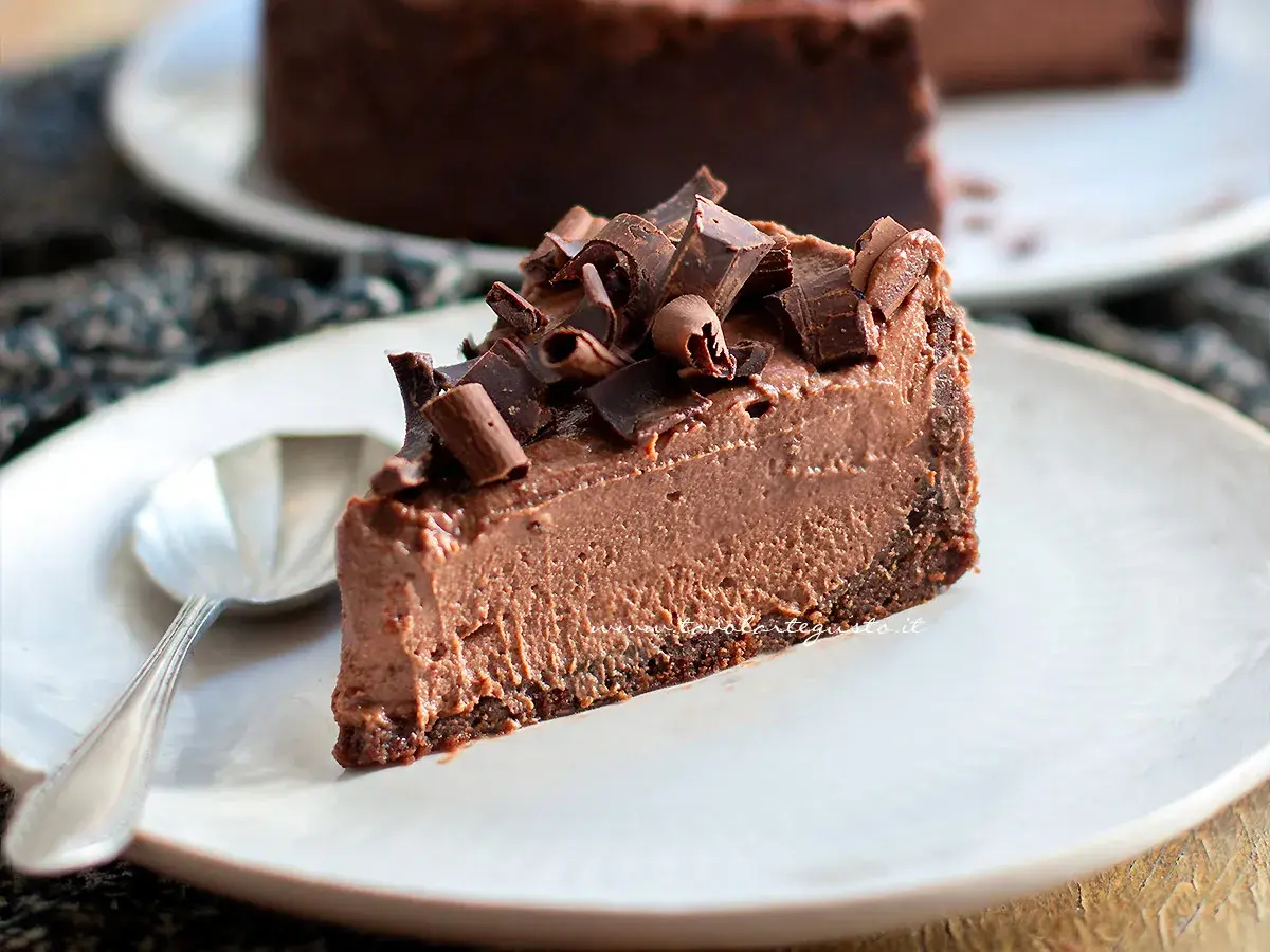 Una fetta di goloso cheesecake al cioccolato, guarnita con riccioli di cioccolato fondente, su un piatto bianco con un cucchiaino.