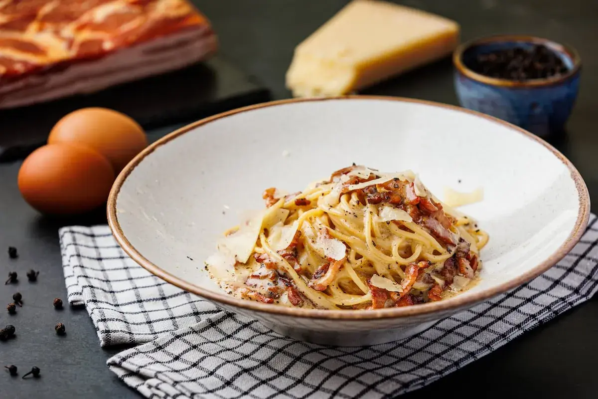 Un plato de spaghetti carbonara cremoso, con trozos de panceta crujiente y queso rallado. Ingredientes frescos como huevos, queso y pimienta negra completan la escena.