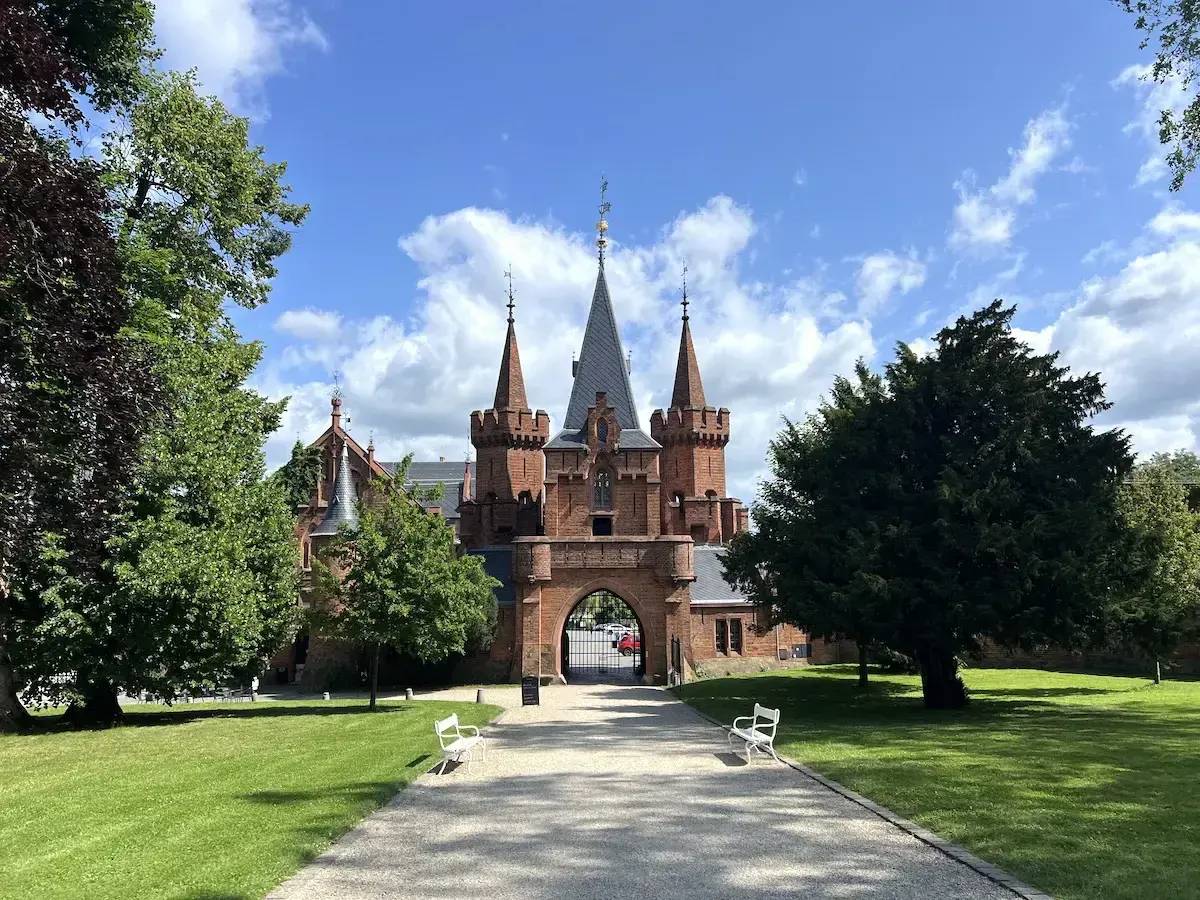 Imponujący, czerwony zamek z wieżyczkami i bramą wjazdową, otoczony zielonym parkiem i drzewami.