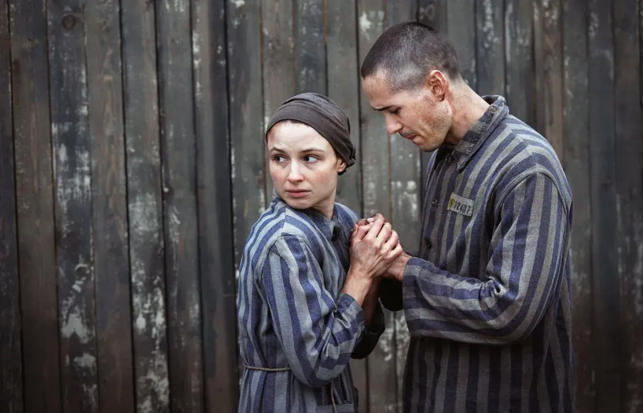 Para więźniów w pasiakach, scena z filmu o Auschwitz. Trzymają się za ręce, patrząc na siebie z nadzieją i strachem.