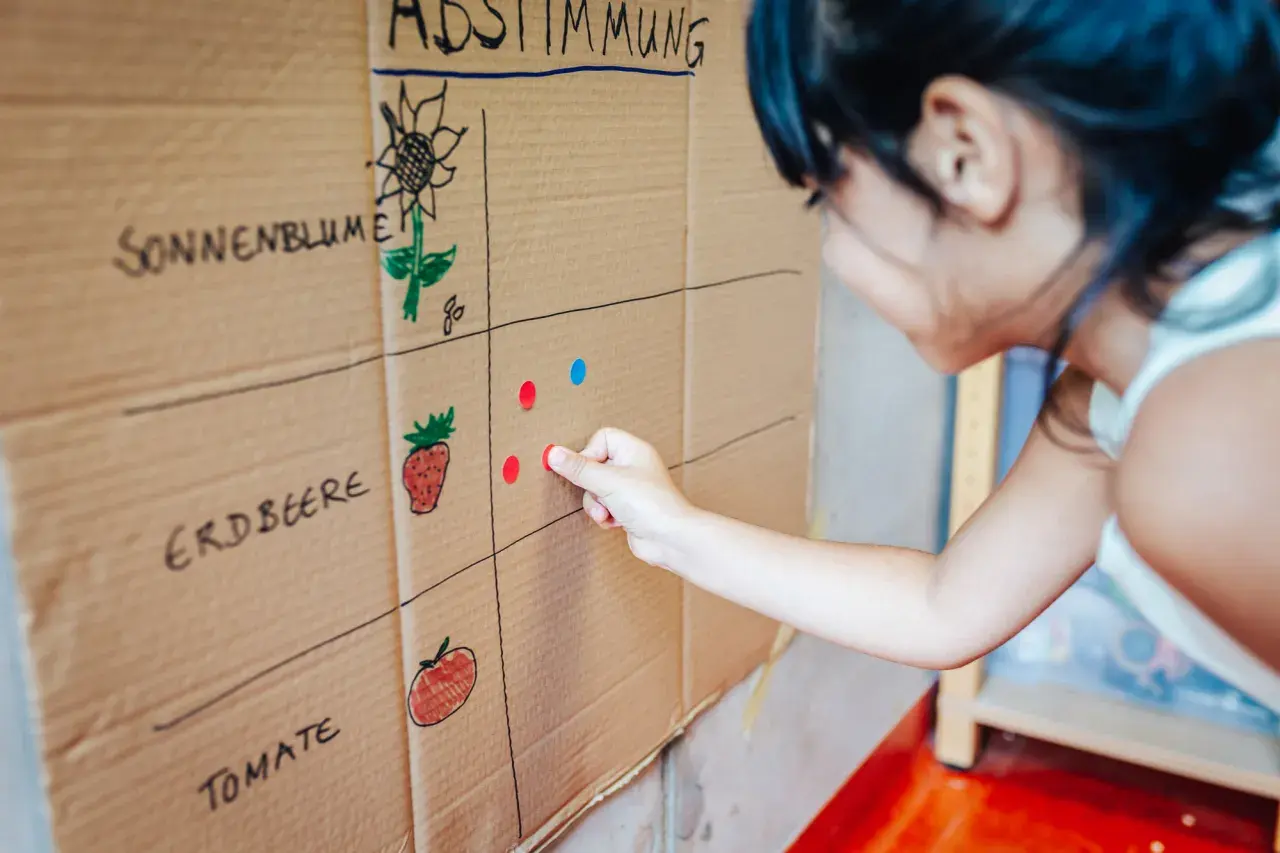 Kind übt Demokratie im Kindergarten: Es wählt per Klebepunkt seine Lieblingsfrucht auf einer selbstgemalten Abstimmungstafel.