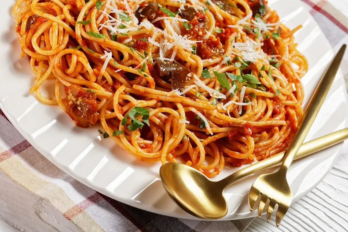 Spaghetti z bakłażanem i pomidorami, polane bogatym sosem sycylijskim, posypane parmezanem i natką pietruszki. Złote sztućce czekają.