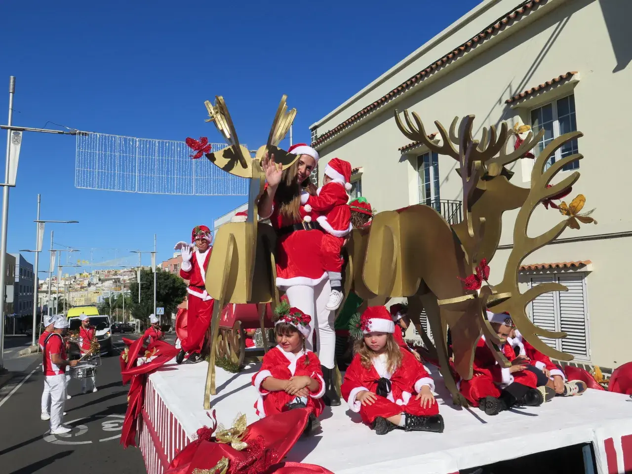 Niños y adultos disfrazados de Papá Noel y elfos participan en una cabalgata de Papá Noel, con un gran reno dorado y decoraciones navideñas.