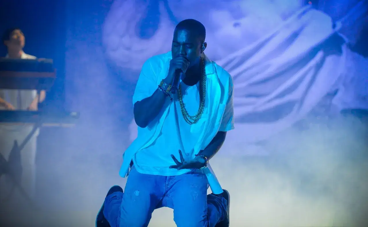 Kanye West na koncercie, klęczy z mikrofonem, otoczony niebieskim światłem i mgłą.
