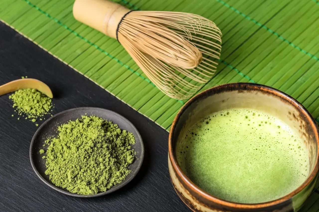 Matcha-Pulver auf einem Teller, daneben eine Schale mit schaumiger Matcha-Creme und ein Bambusbesen.