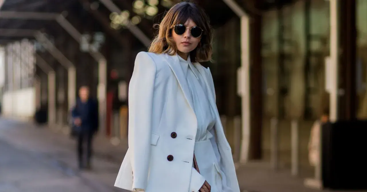 Mujer con traje blanco, gafas de sol redondas y cabello castaño. Su atuendo, elegante y sencillo, refleja el **minimalista significado** de la moda.