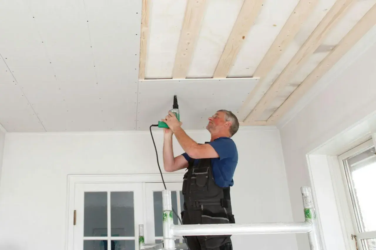 Man på byggställning skruvar gipsskivor i taket. Taket är under renovering med synliga träreglar.