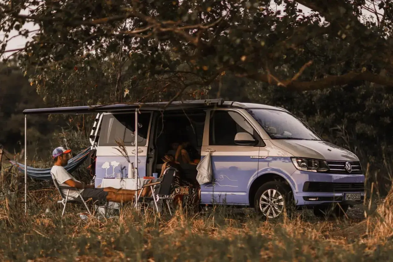 Un homme se détend près d'un camping-car sauvage, avec une table et des chaises installées à l'extérieur. Le véhicule est garé dans un cadre naturel verdoyant.