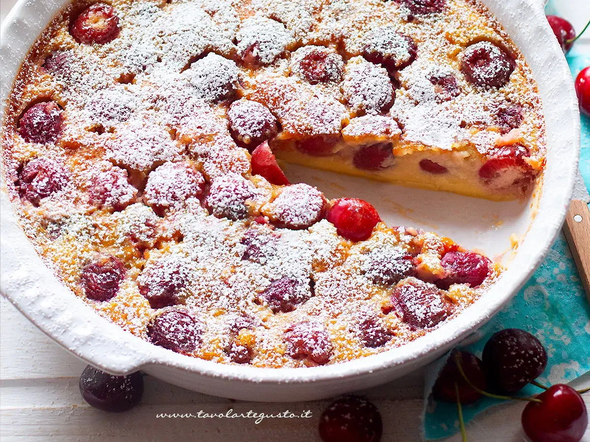Clafoutis di ciliegie appena sfornato, cosparso di zucchero a velo, con una fetta mancante che rivela la morbida consistenza.