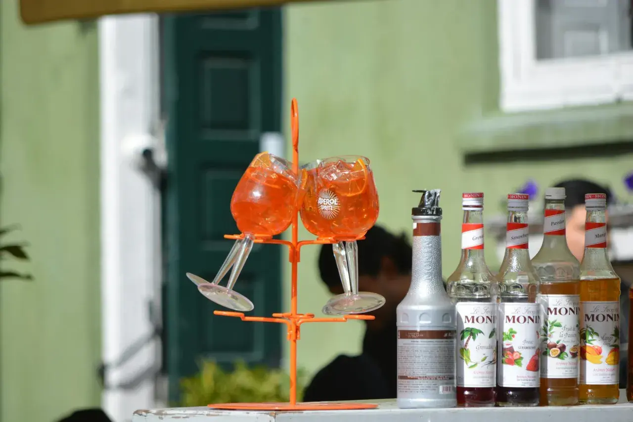 Dwa kieliszki z drinkiem Aperol Spritz na pomarańczowym stojaku, obok butelki syropów Monin.