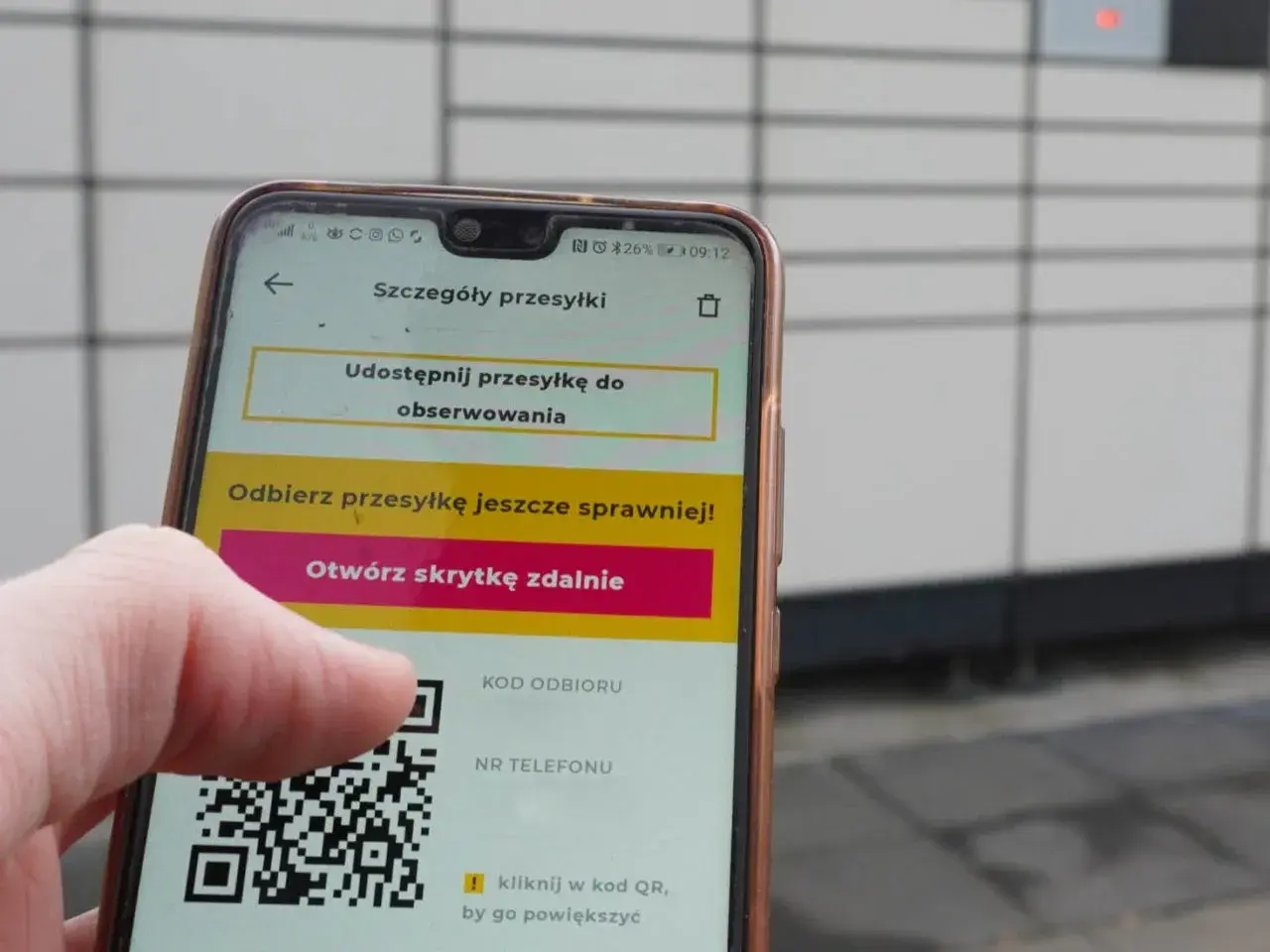 Dłoń trzyma smartfon z aplikacją InPost, wyświetlającą kod QR i opcję otwarcia skrytki.