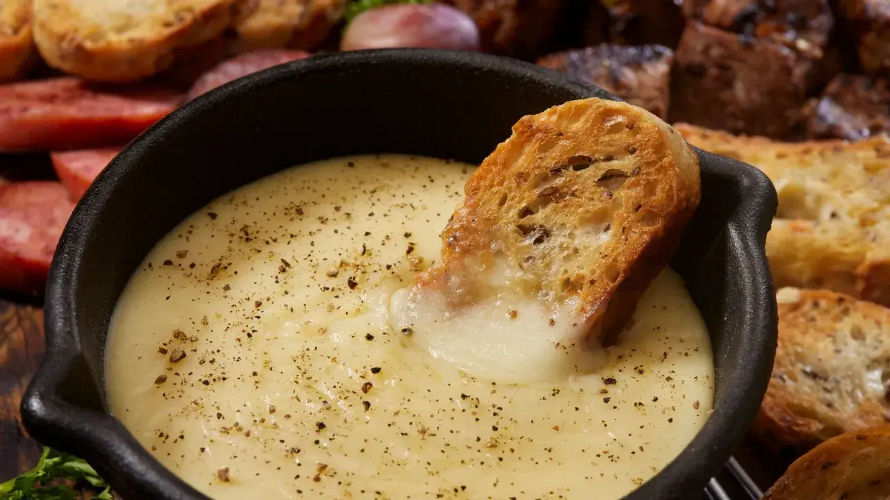 Una fetta di pane croccante si tuffa in una cremosa fonduta di formaggio, cosparsa di pepe nero. Sullo sfondo, salumi e pane tostato invitano a un convivio.