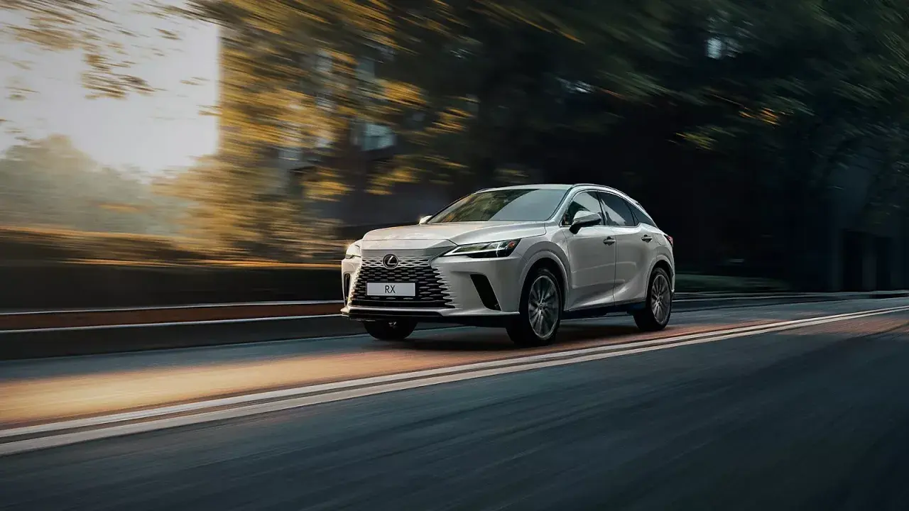 Biały Lexus RX 350h pędzi po drodze, otoczony rozmazanymi drzewami i światłem zachodzącego słońca.