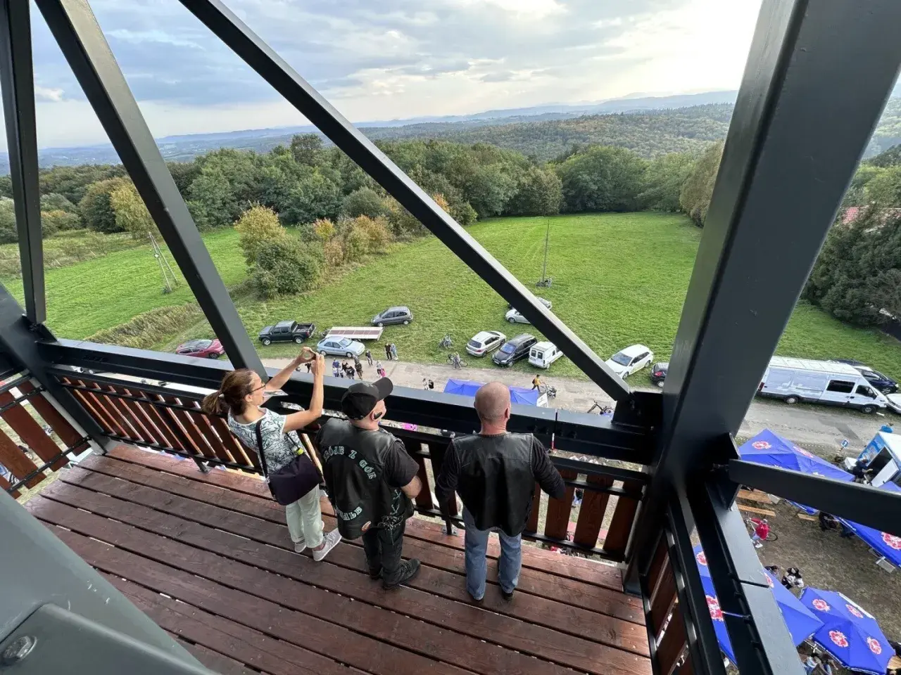 Widok z wieży widokowej na malowniczy krajobraz z lasami i polami. Na dole widać zaparkowane samochody, namioty i ludzi, jakby szykowali się na festyn lub zlot. Może to być nawet zlot motocyklowy, bo jeden z panów ma na sobie kamizelkę z napisem "GÓRY ...