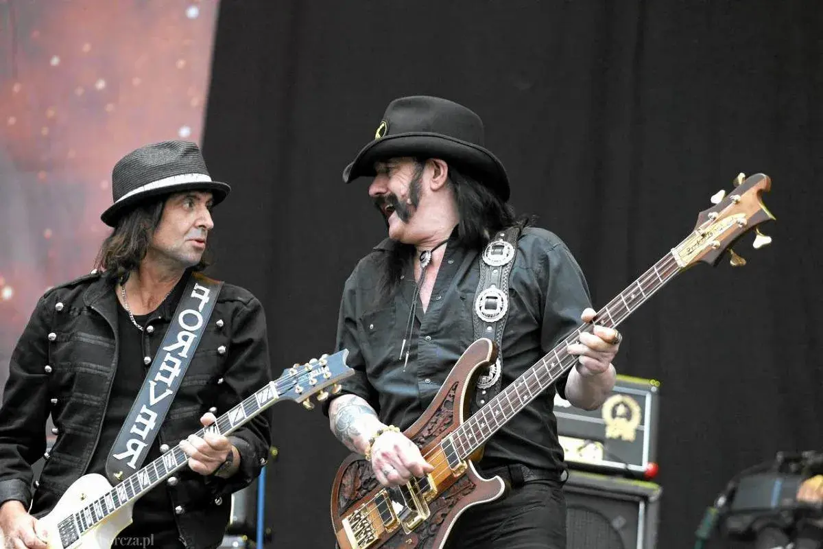 Lemmy Kilmister z zespołem na scenie. Dwóch muzyków gra na gitarach, jeden w kapeluszu, drugi w kowbojskim.