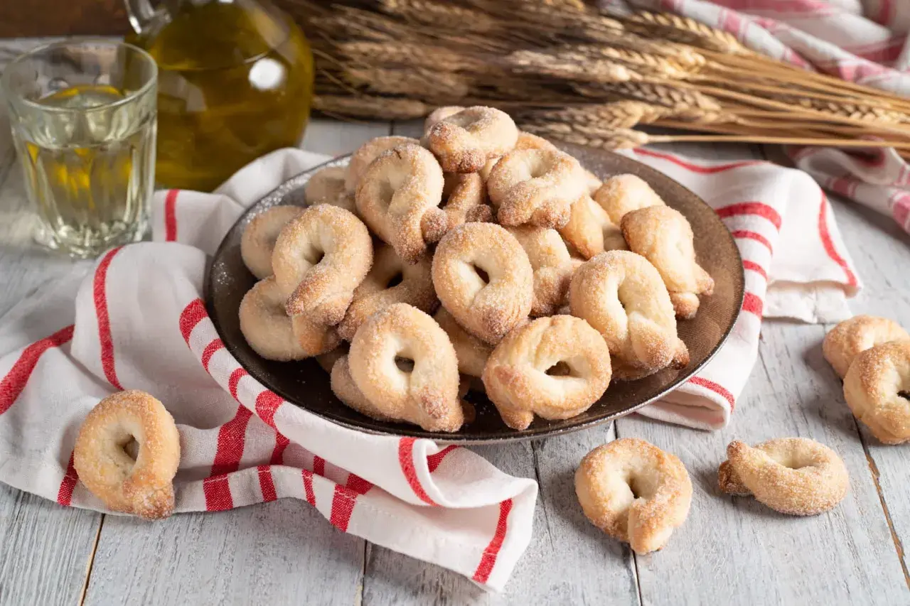 Piatto pieno di taralli dolci al vino bianco, cosparsi di zucchero. Accanto, un bicchiere di vino bianco e una bottiglia d'olio.