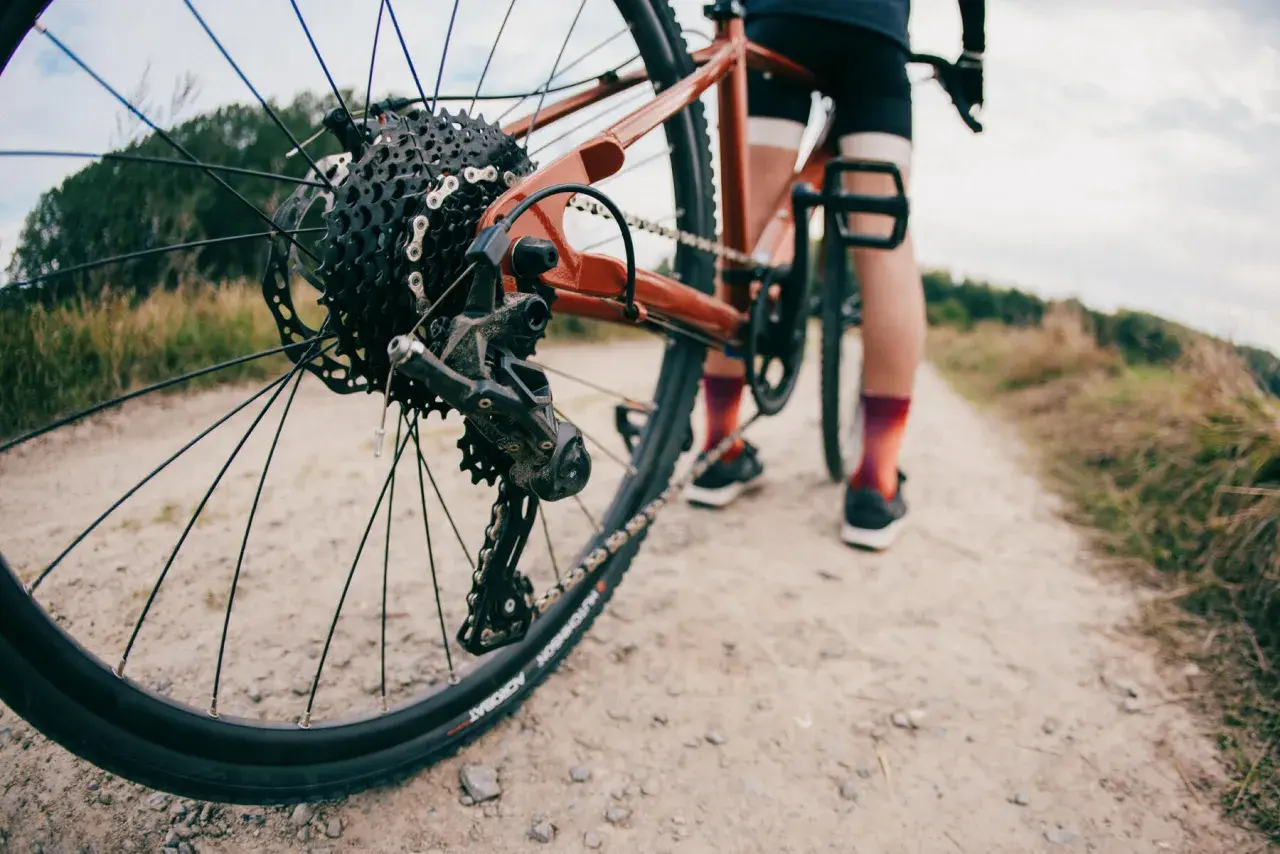 Widok z bliska tylnej przerzutki rowerowej, która wygląda na Shimano Acera lub Alivio, na tle szutrowej drogi i rowerzysty.