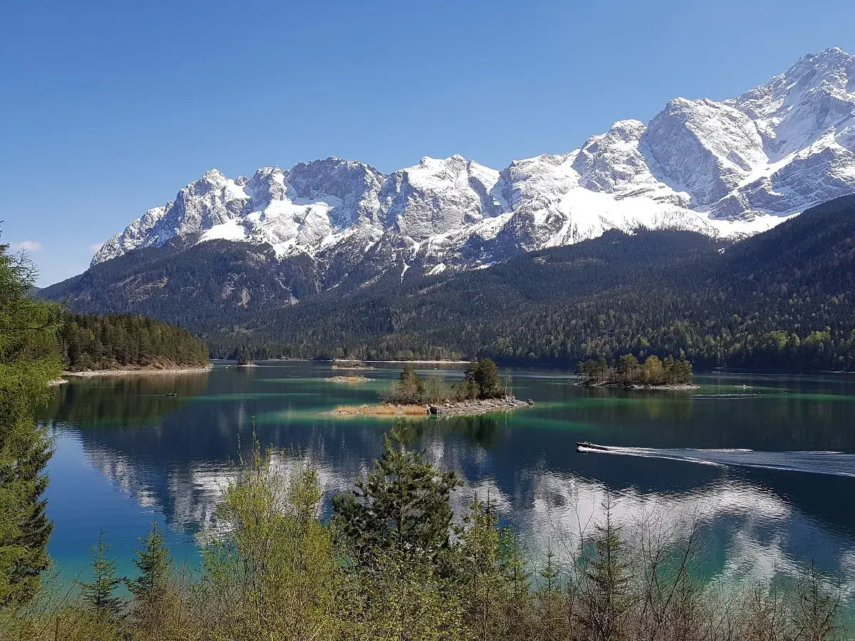 Idyllische Bergkulisse mit schneebedeckten Gipfeln, einem türkisfarbenen See und kleinen Inseln. Ein Boot gleitet über das Wasser, ideal für den Kramerplateauweg Rundweg.