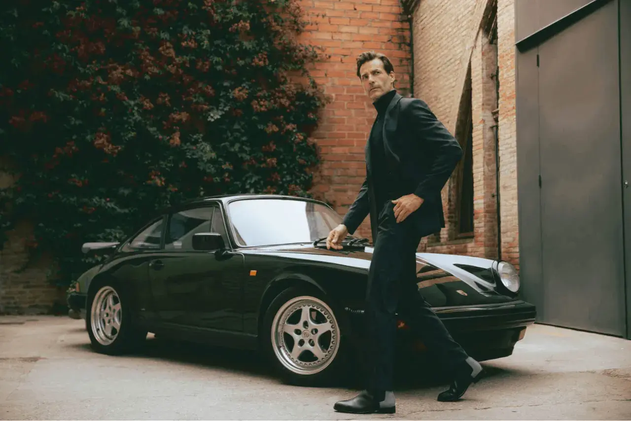 Mann im Anzug und Chelsea Boots lehnt an einem Porsche.