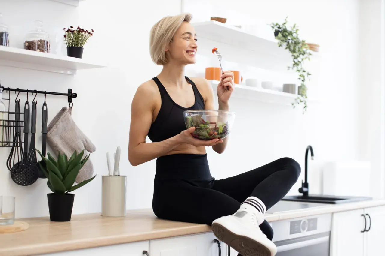 Une femme souriante en tenue de sport mange une salade fraîche. Elle savoure son repas sain dans une cuisine lumineuse.