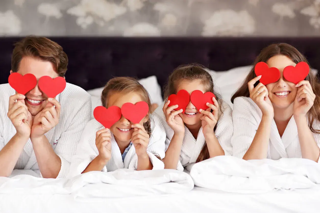 Familia feliz celebra San Valentín para niños, cubriendo sus ojos con corazones rojos en la cama.