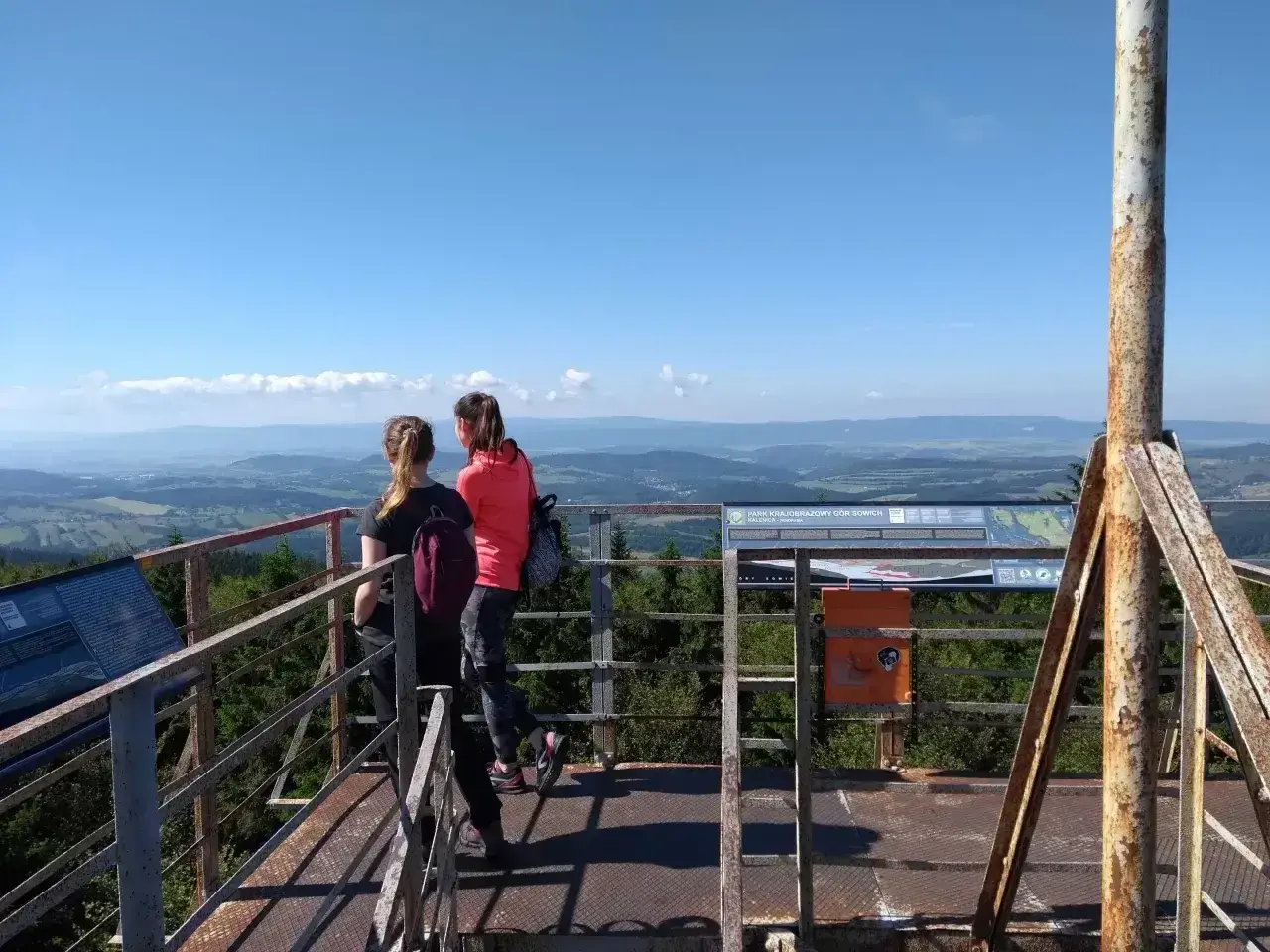 Dwie dziewczyny podziwiają panoramę z wieży widokowej na **kalenicy szczyt**. W tle rozciąga się malowniczy krajobraz Gór Sowich.