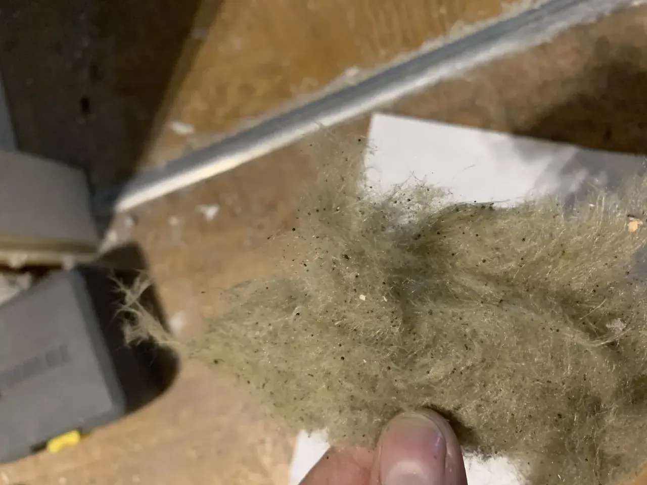 En hand håller en tuss av stenull eller glasull, som är dammig och innehåller små svarta prickar.