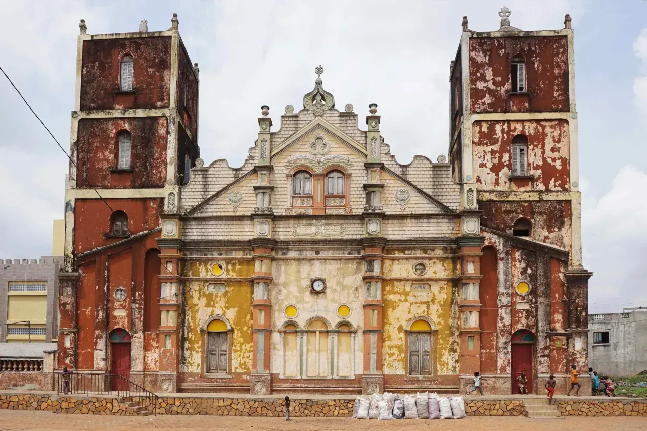 Façade d'une église à Porto-Novo, aux tours jumelles imposantes et à la peinture écaillée, avec des enfants jouant devant.