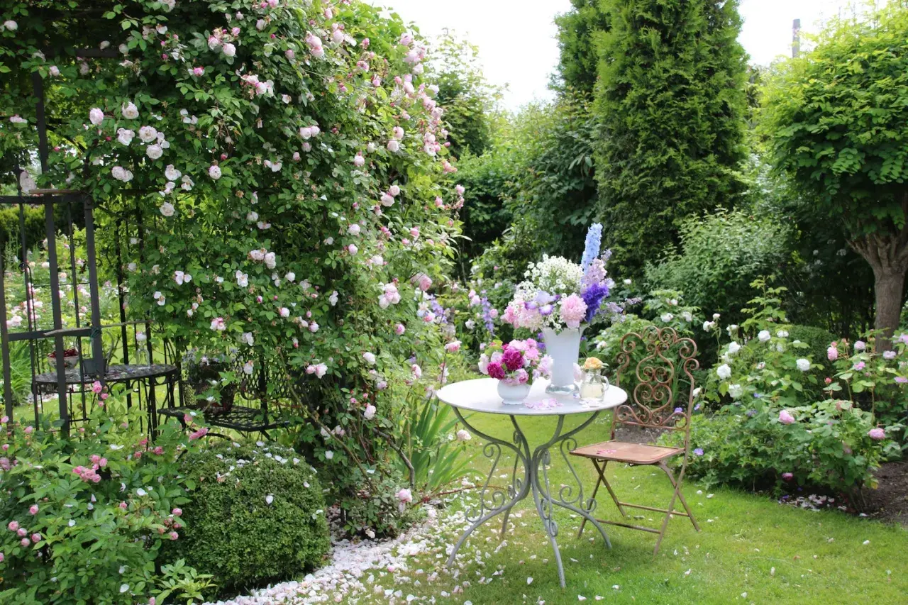 Ein romantischer Garten mit blühenden Rosen, ein Tisch mit Blumenarrangements und ein schmiedeeiserner Stuhl laden zum Verweilen ein. Ein idyllischer Ort, um einen **beet anlegen**-Traum zu verwirklichen.