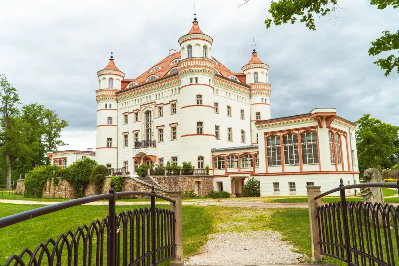Zamek w Łomnicy, biała budowla z czerwonymi dachami i wieżyczkami, otoczona zielenią.