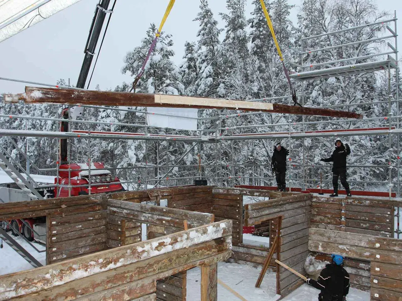 Kranen lyfter timmer för att flytta timmerhus i snöig skogsmiljö. Byggnadsarbetare övervakar arbetet.