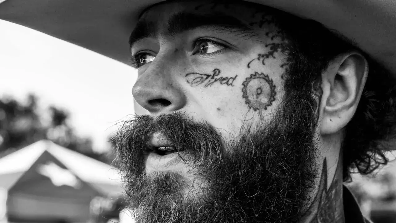 Zbliżenie na twarz Post Malone'a w czerni i bieli, z tatuażem "Tired" na policzku. Fani w Polsce czekają na jego koncert.