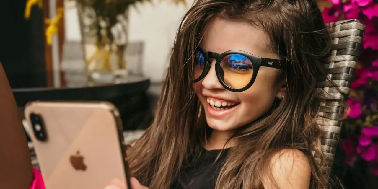 Uśmiechnięta dziewczynka w okularach z filtrem światła niebieskiego patrzy na telefon.