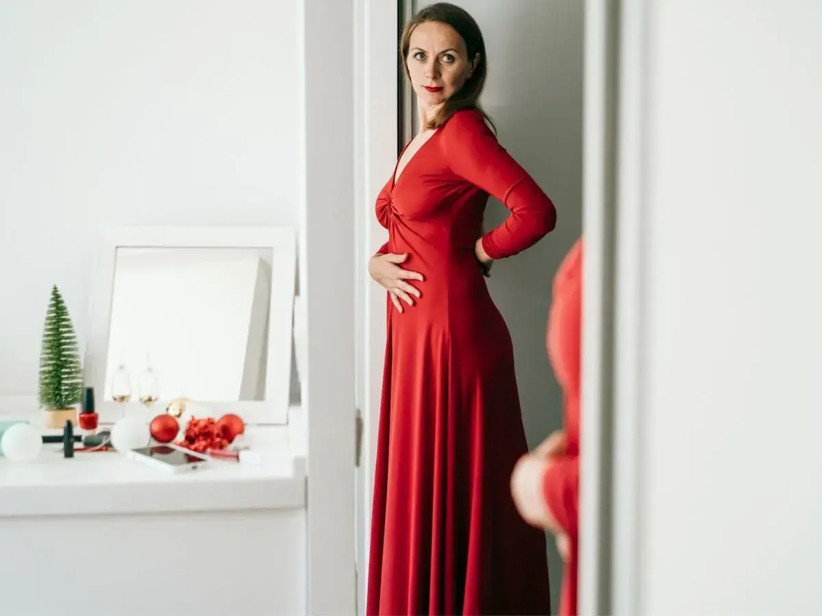 Kobieta w czerwonej, eleganckiej sukience, która optycznie wyszczupla. Na toaletce świąteczne ozdoby i kosmetyki.