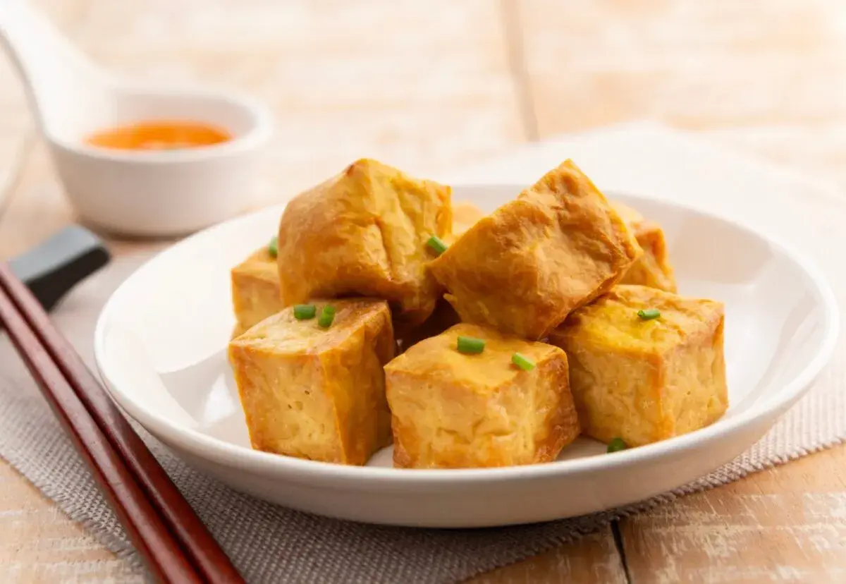 Tofu crujiente dorado, cortado en cubos y adornado con cebollín, servido en un plato blanco con salsa y palillos.