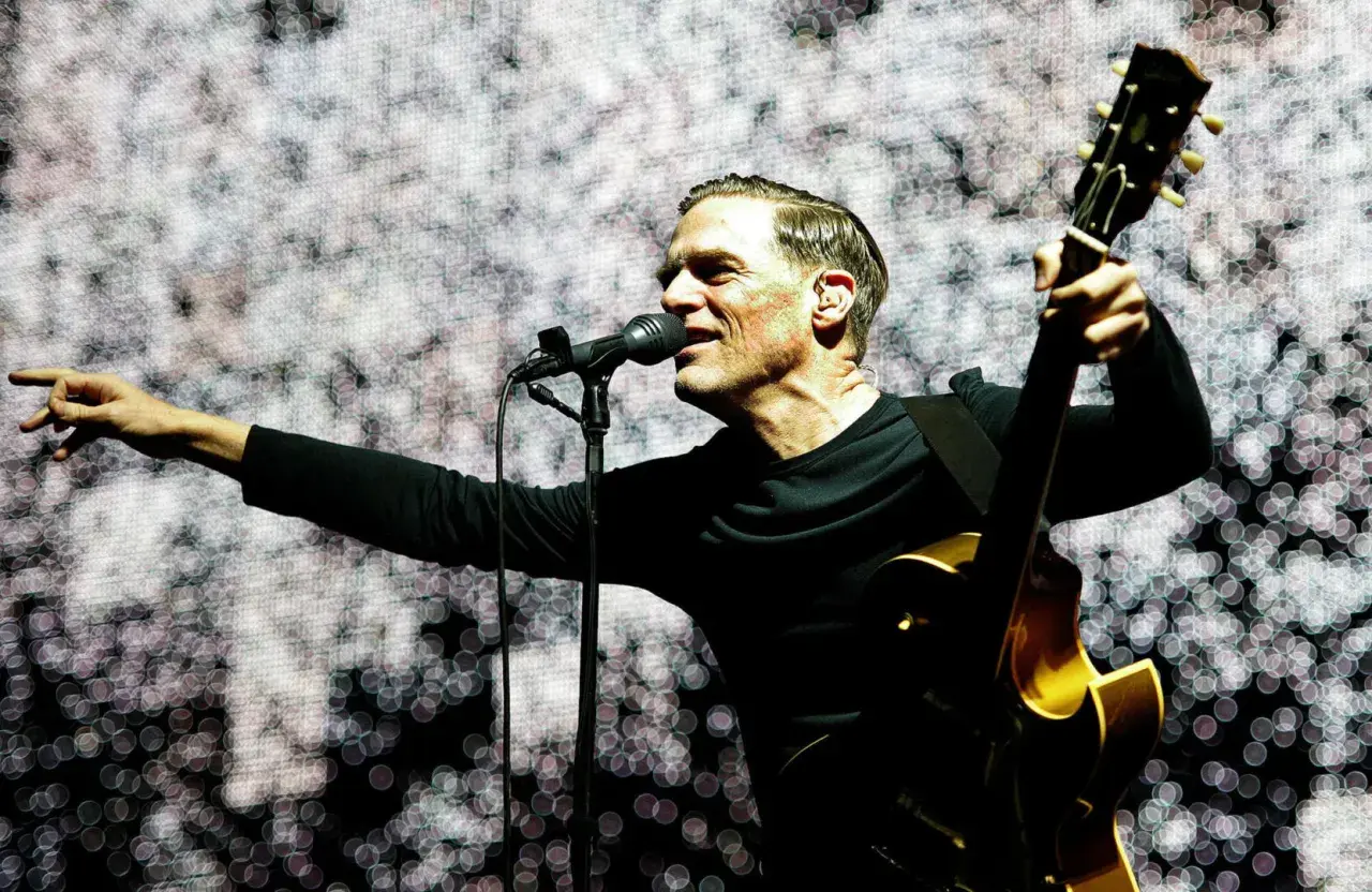 Bryan Adams na koncercie, śpiewa do mikrofonu i gra na gitarze.