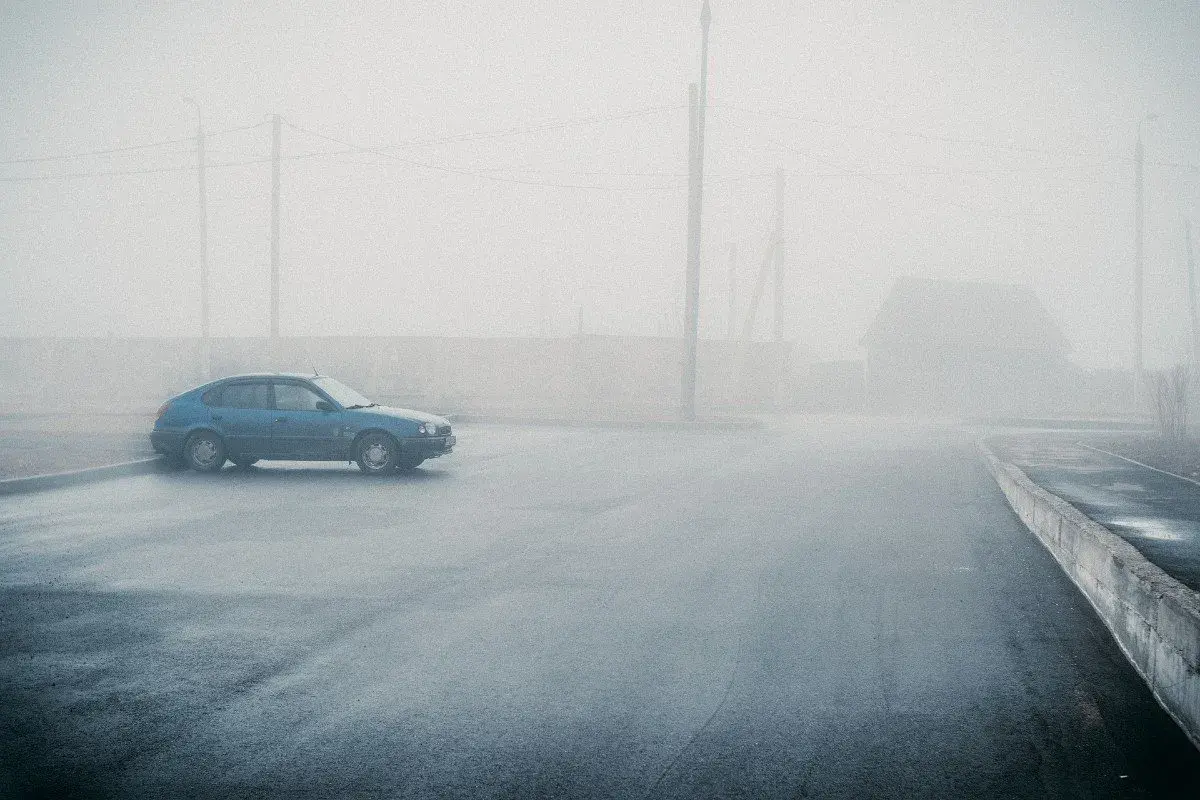 Un coche azul solitario se detiene en medio de una **niebla espesa** que oculta un camino y una casa.