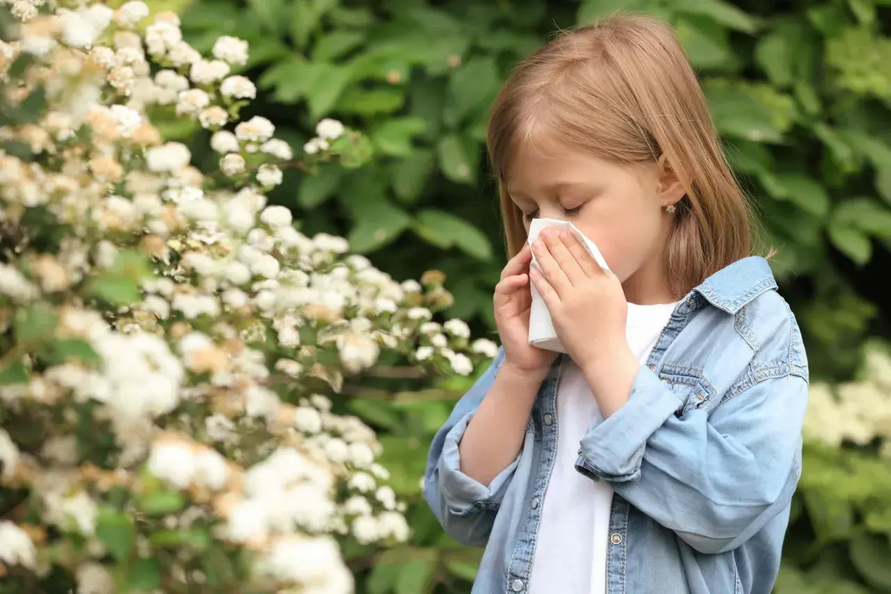 Niña con pañuelo en la nariz, rodeada de flores. Posiblemente sufre de alergia en niños.