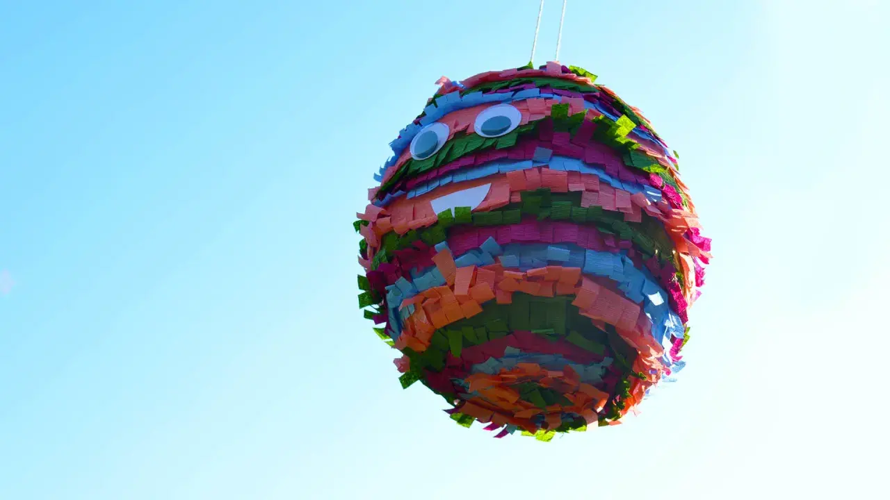 Piñata casera colorida con ojos saltones y boca sonriente, colgada contra un cielo azul claro.